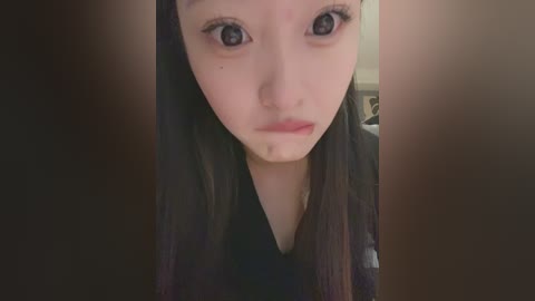 Media: Video of a young Asian woman with fair skin and long black hair, wearing a black top. She has large, expressive eyes and a neutral expression. Background is blurred, suggesting an indoor setting.