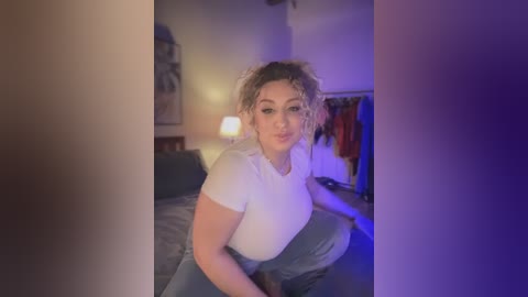 Video of a curvy woman with curly blonde hair, wearing a tight white shirt and blue jeans, sitting on a bed in a dimly lit bedroom with purple lighting.