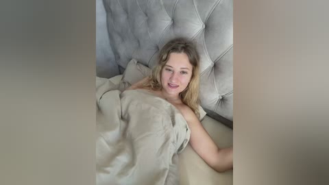 Video of a smiling, light-skinned woman with wavy blonde hair, partially covered by a beige blanket, lying on a light-colored bed with a tufted, gray headboard.