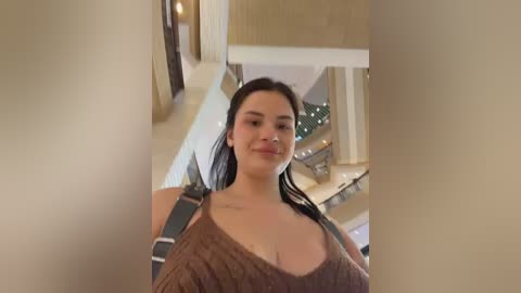 Video of a smiling young woman with long black hair, wearing a brown, knitted sweater, standing in a modern, beige-toned, indoor space with a spiral staircase in the background.