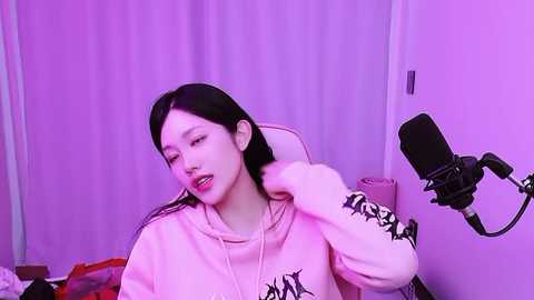 Video of a young Asian woman with long black hair, wearing a pink hoodie, sitting in a white chair, singing into a black microphone. Purple lighting, casual setting, soft focus.