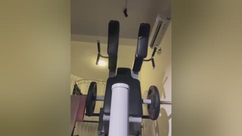 Video of a home gym with a black and white adjustable bench, dumbbells, and a ceiling fan. The walls are painted yellow, and a white air conditioning unit is mounted on the wall.