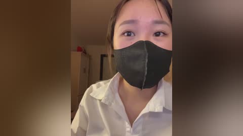 Video of a young East Asian woman with fair skin, wearing a black face mask and a white button-up shirt. The background shows a dimly lit indoor setting with beige walls and a wooden cabinet.