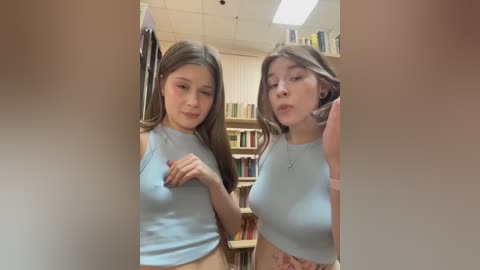 Media: Video of two young women with long hair, wearing light blue tank tops, standing in a book-filled room, both looking down, with one touching her breast.