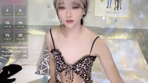 A video of a slender, young Asian woman with light skin and shoulder-length, blonde hair, wearing a leopard-print bra. She sits on a bed with white sheets, in a dimly lit bedroom.