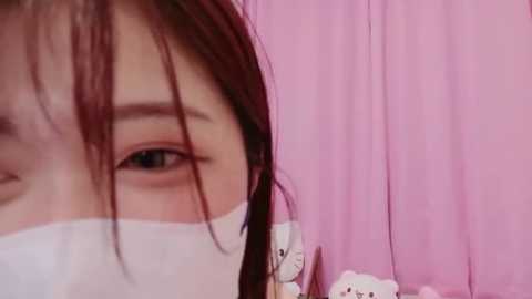 Media: Video of a young woman with light skin, brown hair, and blue eyes, partially obscured by a white surgical mask, against a soft pink background.