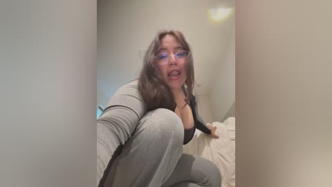 Video of a plus-sized woman with long brown hair, wearing glasses and a tight gray top, squatting provocatively in a bedroom.