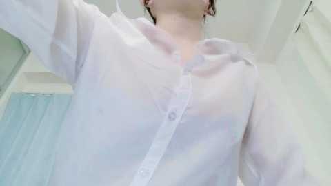 Media: Video of a woman wearing a see-through white button-up shirt, revealing her bra underneath. Her fair skin and brown hair are visible. Background shows a white wall and a blue shower curtain.