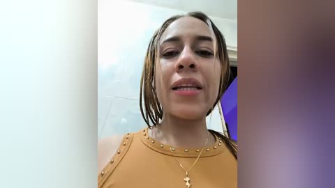 Video of a young, light-skinned woman with wet, shoulder-length brown hair, wearing a mustard-yellow blouse with gold studs, smiling slightly, in a bathroom with white tiles and a blue towel.