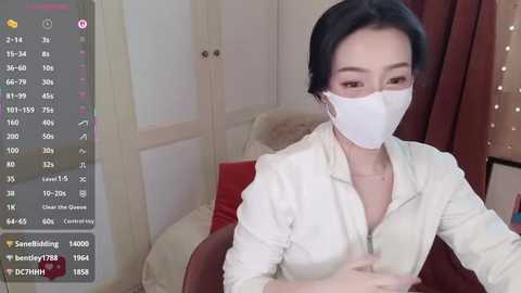 Video of a woman with short black hair, wearing a white mask, blouse, and red cushioned chair. Background shows white doors and beige curtains.