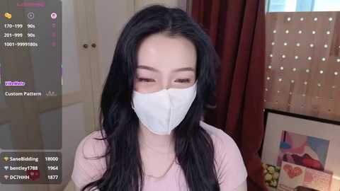 A video of an Asian woman with long black hair, wearing a white face mask, pink top, and a necklace, indoors with red curtains and a fruit basket in the background.