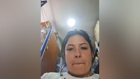 Media: Video of a woman with dark hair and brown skin, wearing a white shirt, standing in a cluttered room with wooden cabinets and hanging items, partially out of focus.