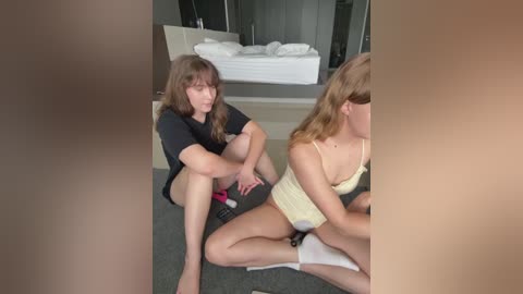 A video of two young women in a modern bedroom. One wears a black shirt, pink shorts, and sits on a gray carpet. The other, in a yellow tank top, sits on a white towel, facing away.