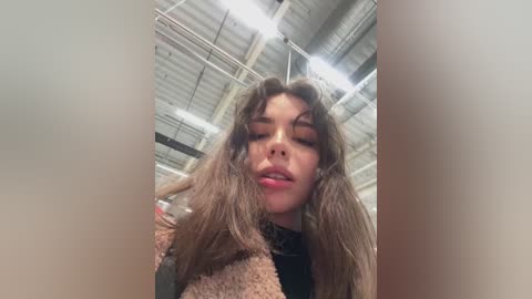 Media: A young woman with long, wavy brown hair, wearing a black top and a light beige coat, poses indoors with a ceiling of exposed metal beams and fluorescent lights.