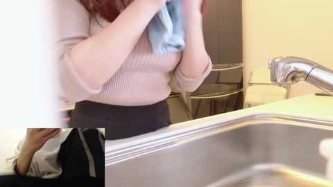 A video captures a woman in a bathroom, holding a blue towel, wearing a beige sweater and black pants. The sink and mirror are visible in the background.