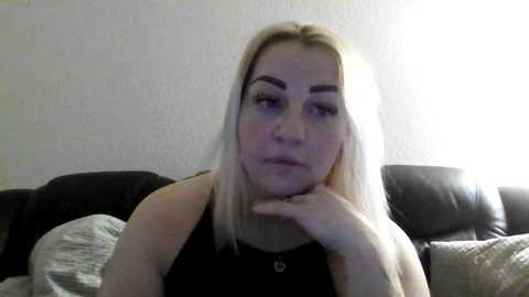 Video of a fair-skinned, blonde woman with straight hair, resting her chin on her hand, wearing a black top, seated on a gray couch against a plain white wall.