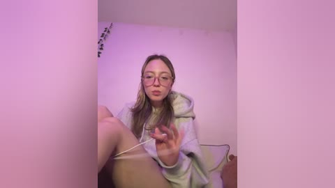 Video of a young, fair-skinned woman with glasses, light brown hair, wearing a gray hoodie, sitting on a beige couch in a dimly lit room, holding a phone, and making a playful expression.