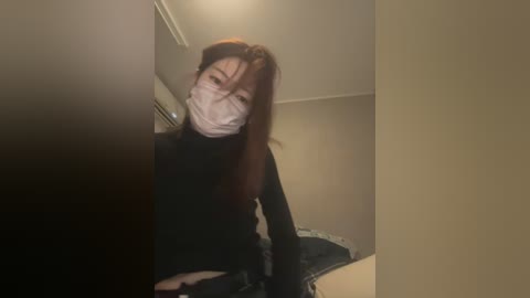 Media: Video of a woman with long, reddish-brown hair wearing a black turtleneck and a white mask, standing in a dimly lit room with beige walls.