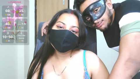 Video of a young woman with long black hair wearing a black mask and light blue top, seated beside a man in a black mask and striped shirt, in a room with a digital thermometer screen.