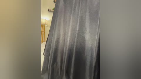 A video shows a grey, silky curtain partially covering a beige wall in a dimly lit room. A white lamp with a wooden base is visible in the background.