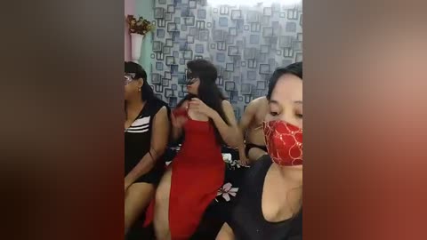 Video of three Asian women in a dimly lit room with a patterned backdrop; one in a red dress, another in a black outfit, and a third with a red mask.