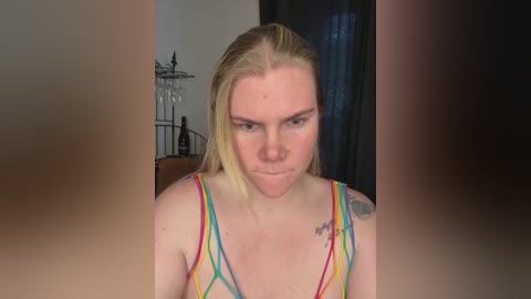 A video of a blonde woman with a stern expression, wearing a colorful, multi-striped tank top, with a tattoo on her right shoulder. Background shows a dark curtain and a metal birdcage.
