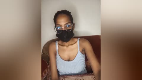 Video of a young Black woman with medium brown skin, wearing a light blue ribbed tank top, black mask, and glasses, sitting on a patterned couch.