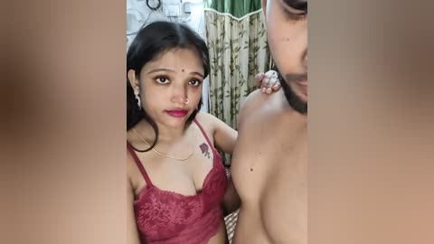 Video of a young woman with dark skin, medium build, and long black hair, wearing a red lace bra, posing intimately with a man's bare chest, both in a dimly lit room with green curtains.