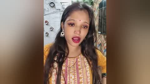 Media: Video of a young South Asian woman with long, dark hair, wearing a mustard-yellow kurta with intricate embroidery, and red bindi, against a blurred, rustic background.