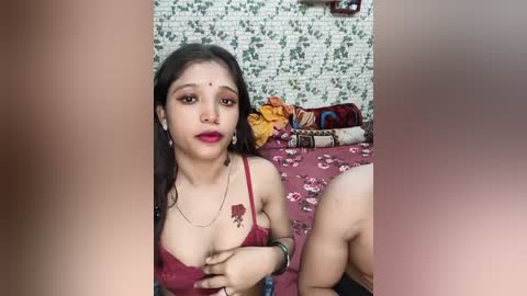 Media: Video of a young South Asian woman with long black hair, wearing a red lace bra and gold jewelry, sitting on a bed with floral sheets in a room with white brick wallpaper.