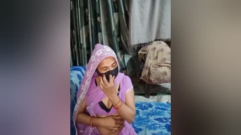 A video shows an Indian woman wearing a purple sari, black mask, and headscarf, seated on a blue-patterned blanket, covering her chest with her hands. The background features green and white curtains and a beige blanket.