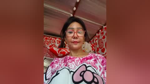 Video of an Asian woman with medium skin tone, dark hair, and red-framed glasses, wearing a pink floral top, lying on a bed with red and white patterned pillows.