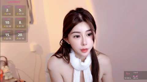 A video of a young East Asian woman with long brown hair, fair skin, and delicate features, wearing a white turtleneck sweater, sitting indoors against a soft beige wall.