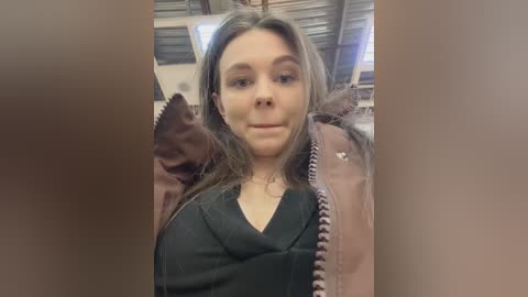 A video of a young woman with long brown hair, wearing a black top and a pink jacket, standing indoors with metal walls and a white ceiling.