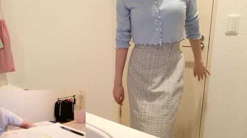 Video of a woman standing in a bathroom, wearing a light blue button-up shirt and a gray plaid pencil skirt. Background includes a white bathtub, beige walls, and a door.