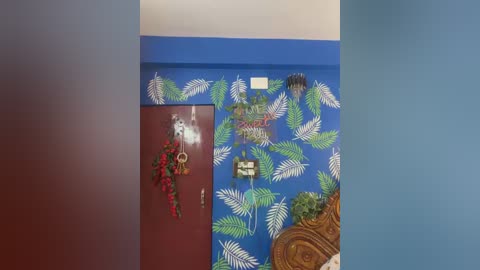 Video of a vibrant room with blue walls adorned with green leaf patterns. A maroon door with a red flower wreath, a wooden headboard, and a potted plant with lush green leaves are visible.