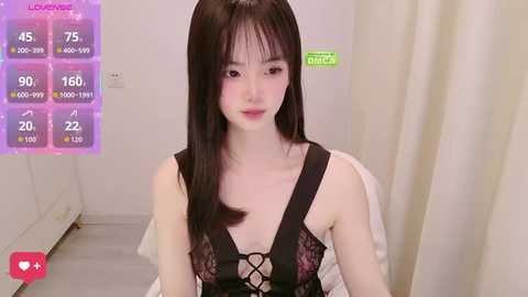 Video of an Asian woman with long black hair, wearing a black lace bralette, sitting on a bed. Background features a white wall and a curtain.