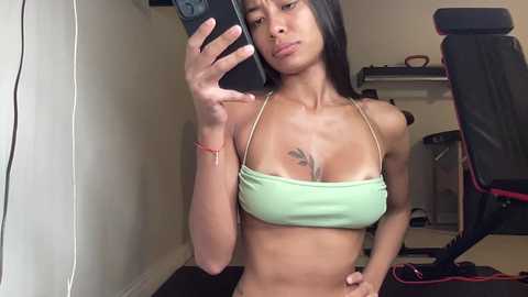 Media: Video of a young, slender Asian woman with long black hair, wearing a light green bikini top, taking a mirror selfie in a dimly lit room with gaming equipment.