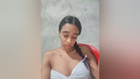 Media: Video of a young woman with medium skin tone, dark hair, and a slender build, wearing a white, off-the-shoulder top and a pink bracelet, sitting on a red chair against a textured gray wall.