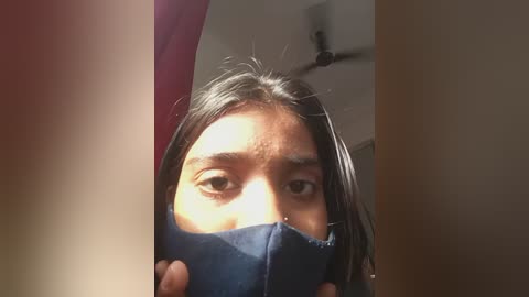Media: A video of a young woman with long black hair, wearing a dark blue face mask, covering her mouth and nose, taken indoors with a ceiling fan and red wall visible in the background.