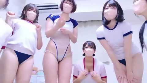 Video of four young Japanese women in white and navy-blue sportswear, wearing face masks, posing in a modern, brightly-lit room.