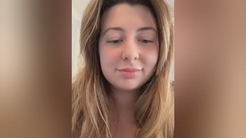 Video of a young woman with long, straight blonde hair, fair skin, and light pink lips, smiling slightly. She is indoors with soft, neutral background lighting.