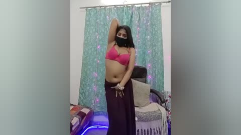 Video of a woman with medium skin tone, wearing a pink bra, black mask, and dark skirt, posing confidently in a dimly lit room with teal curtains and a black chair.