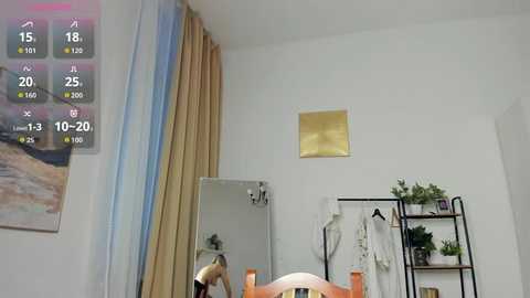 Video of a modern bedroom with beige curtains, a white bed, a black metal shelf with plants, and a digital clock showing 13:30.