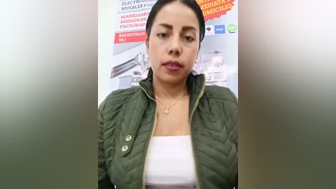 Video of a Latina woman with medium skin tone, dark hair, and a medium build, wearing a green puffer jacket over a white tank top. She stands in front of a colorful, multilingual advertisement background.