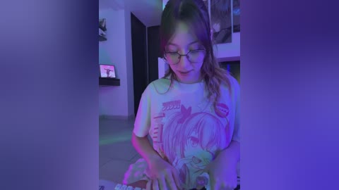 A video of a young girl with glasses and long brown hair, wearing a \"Genshin Impact\" T-shirt, sitting indoors under purple and green lighting.