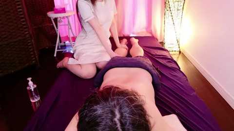 A video of an Asian woman in a white dress massaging an Asian man lying face down on a purple mat in a dimly lit room with pink and purple curtains and a wicker chair.