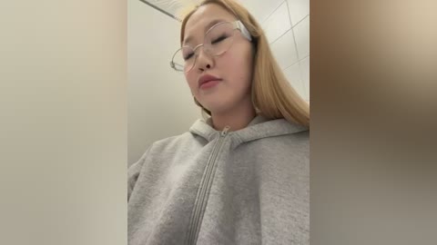 A video of an Asian woman with long blonde hair and fair skin, wearing glasses and a grey hoodie, standing in a white-tiled bathroom. Her expression is neutral.