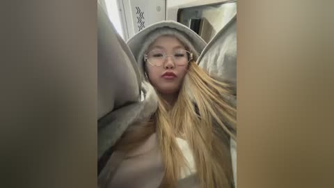 Video of a young Asian woman with long blonde hair and glasses, wrapped in a gray blanket, looking relaxed in a cozy, dimly lit room.