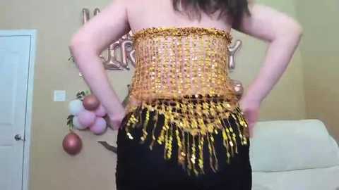 Media: Video of a woman's back, wearing a gold sequined top and black pants. The background features a white door, decorative garland, and a \"Merry\" sign.